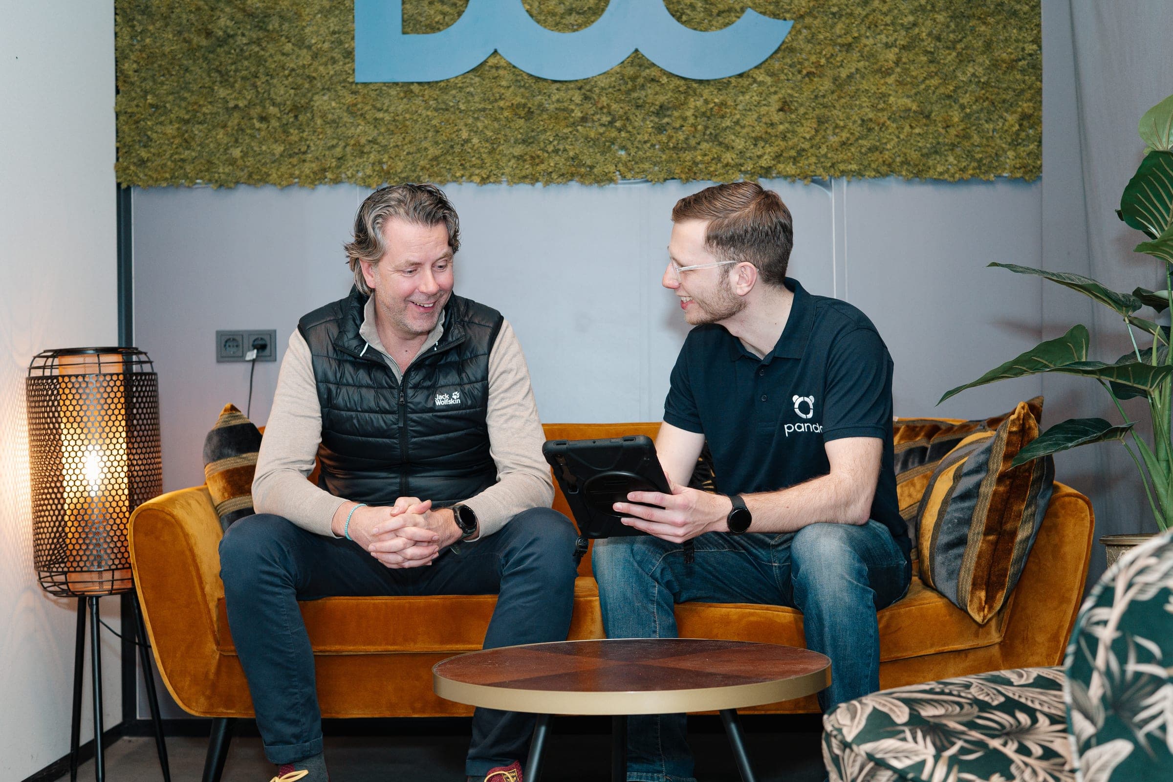 Panda-adviseur in gesprek met klant op gele bank, tablet met portefeuille-data zichtbaar — typisch advies-moment voor maatwerkadvies